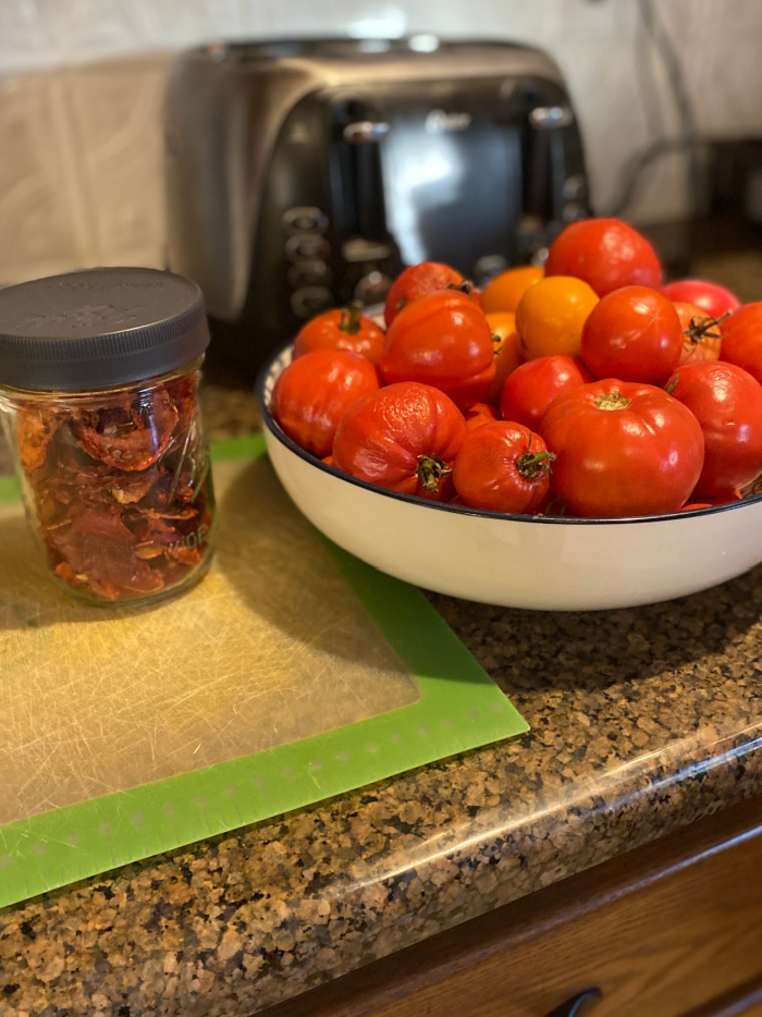 Fresh tomatoes in a bowl on a counter, alongside a jar of dehydrated tomatoes.