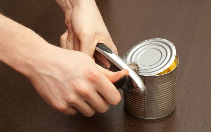 Woman using a manual can opener to open a can of corn.