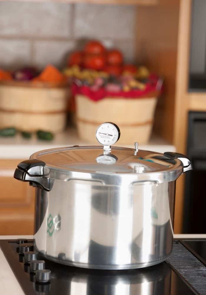 A pressure canner sitting on a stovetop ready to can vegetables, with bushels of vegetables in the background.