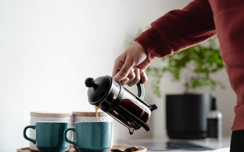 A person pouring coffee from a glass french press into two teal ceramic coffee mugs.