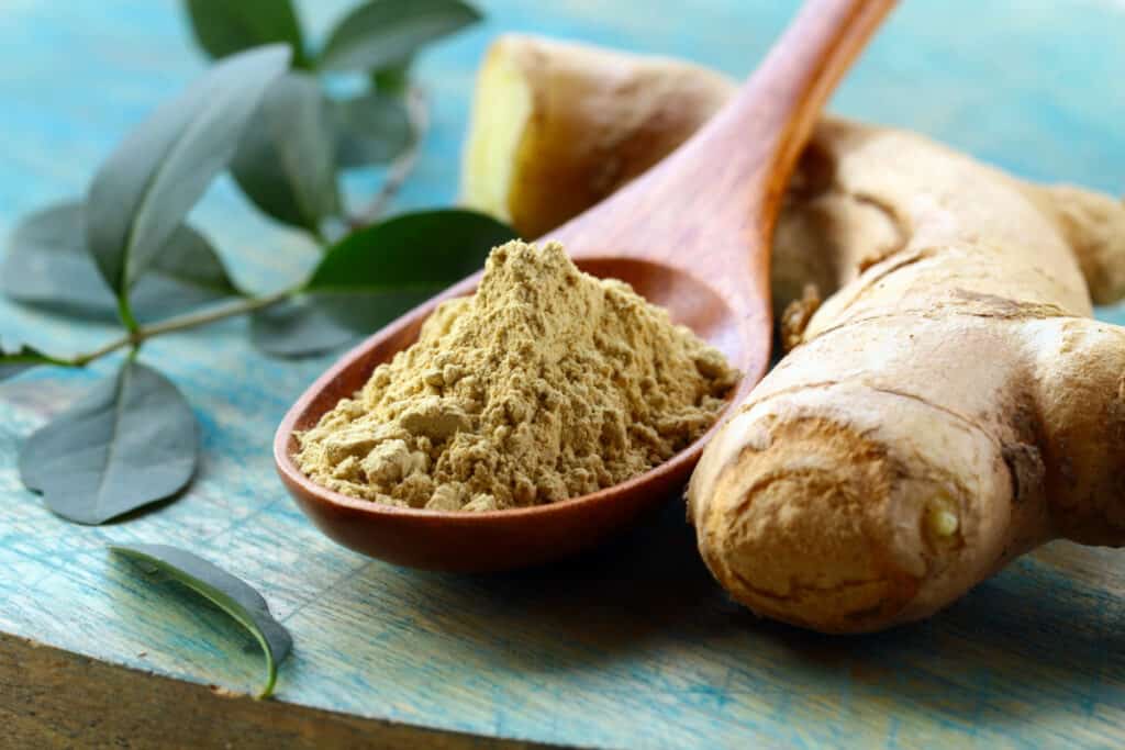 Dried, ground ginger on a wooden spoon beside a piece of ginger root on a wooden table.