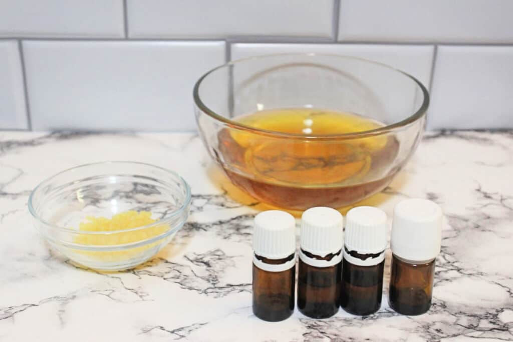 four bottles of essential oil on a marble countertop next to a glass dish of beeswax pellets and a glass bowl of infused oil.
