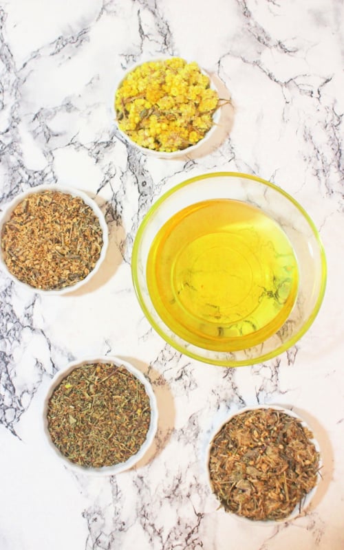 Ingredients for herbal infusion- dried herbs, flowers and avocado oil in individual containers on a marble background.