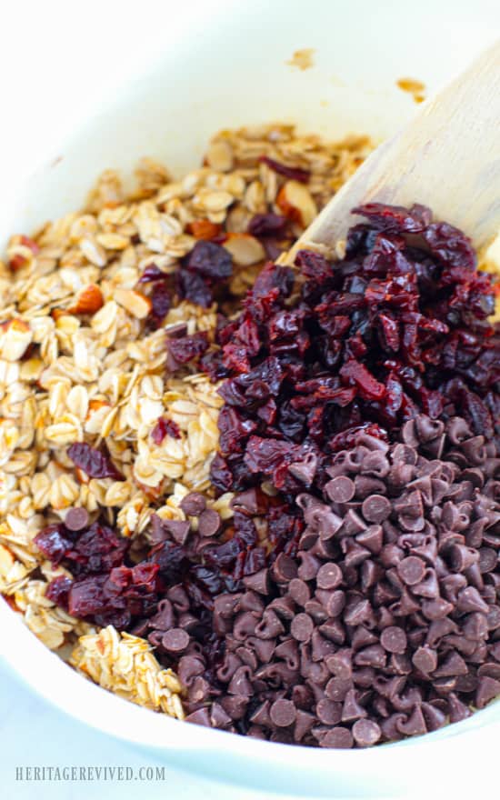 Mixing together chopped cherries, chocolate chips, maple syrup and oats with a wooden spoon in a white mixing bowl. 