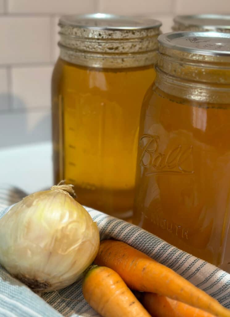 Finished quart jars of homemade chicken broth next to a tea towel with onion and carrots on a countertop.