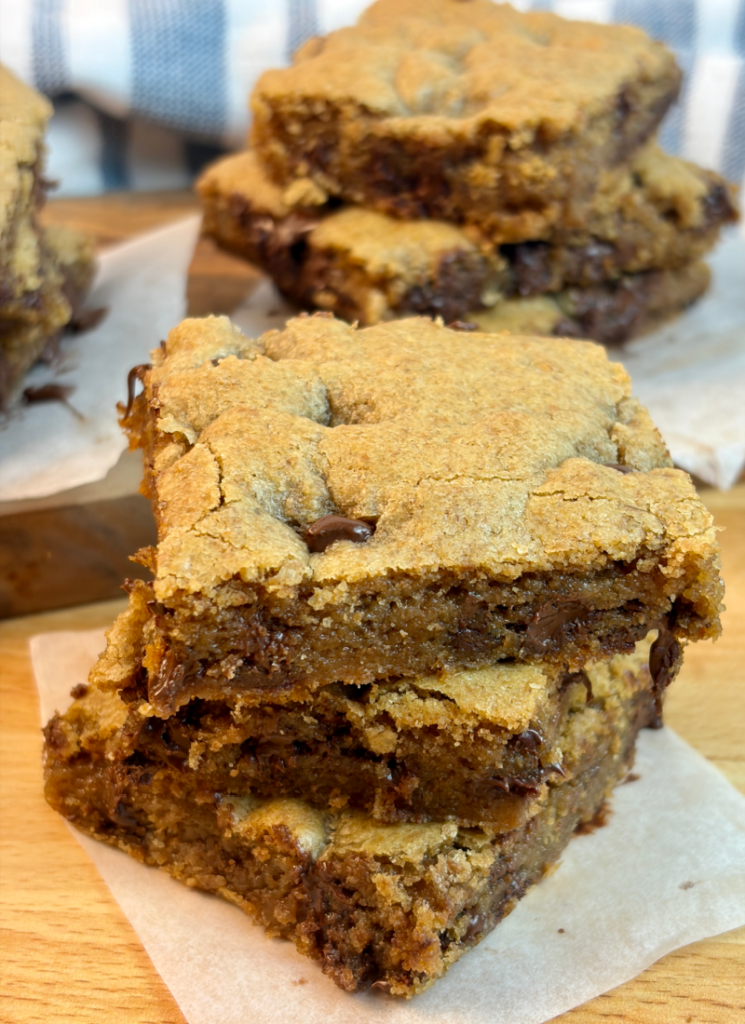 Cookie bars cut into squares and stacked on parchment paper on wooden counter.