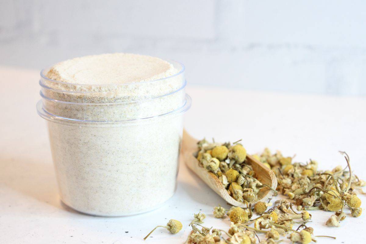 Bath salts in glass mason jar with spoon and dried chamomile on table beside it.