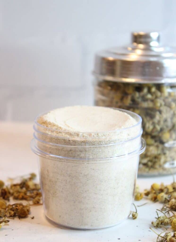 Vertical photo of bath salt mixture in an open mason jar with glass container of chamomile flowers in background and scattered on table.