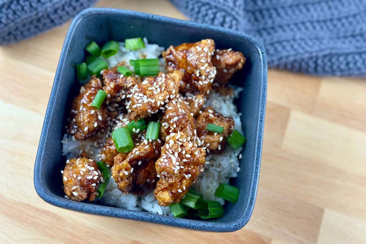 Sesame chicken in square gray bowl on a butcher block countertop.