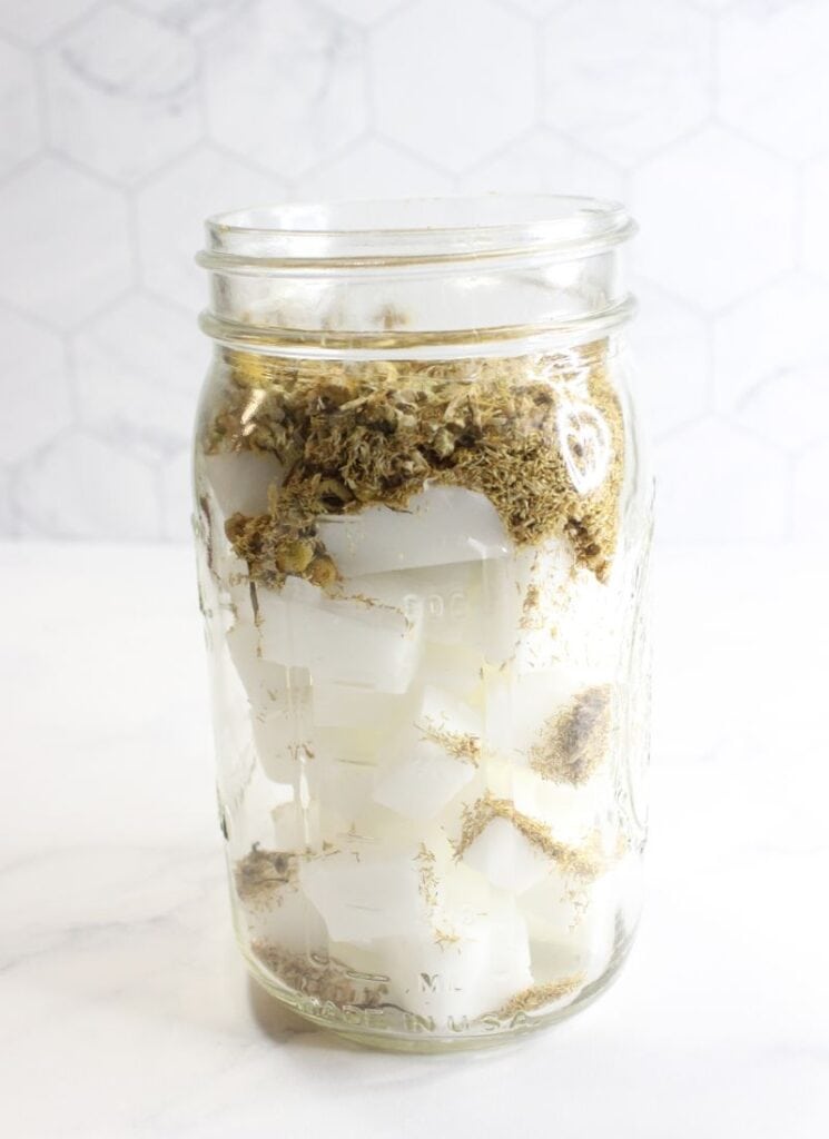 Mason jar filled with soap base and chamomile flowers, ready to melt and infuse over double boiler.
