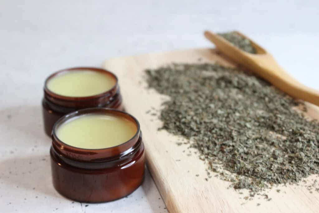 Finished lip balm in two glass jars next to a cutting board with dried lemon balm.