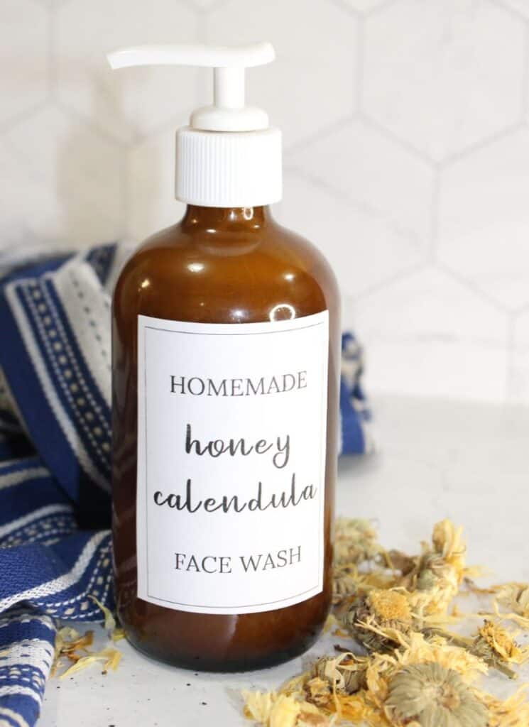 Cleanser in brown pump bottle on counter with calendula flowers scattered on surface and text on bottle that reads "homemade honey calendula face wash"