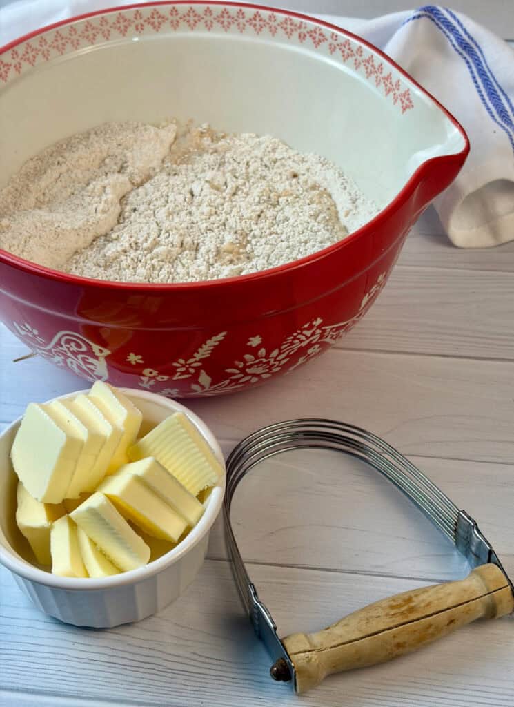 Bowl of dry ingredients with butter in dish beside it and pastry blender on table.