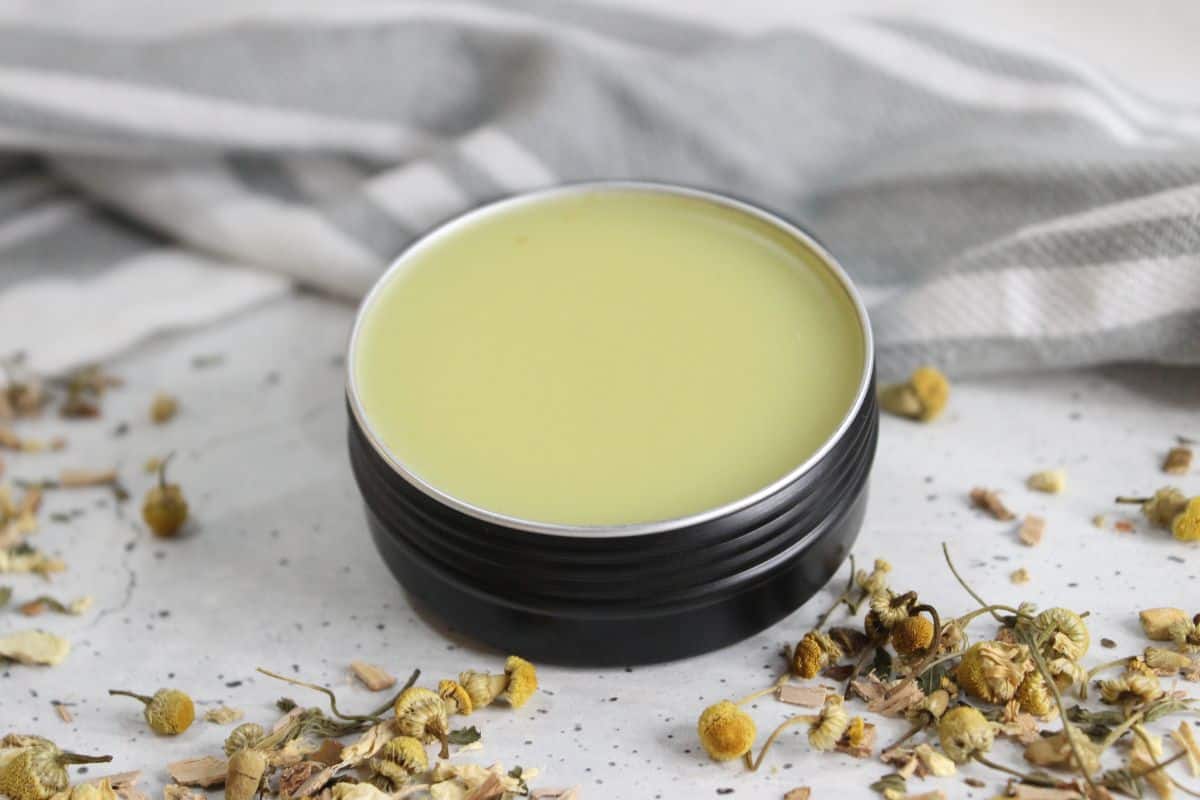 Headache salve in a tin container with herbs and a striped tea towel in background.