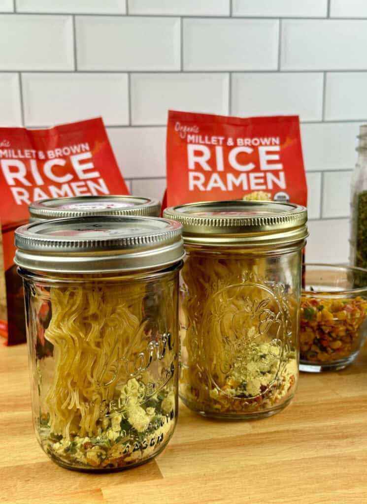 Two jars with rice ramen noodles, seasonings and dehydrated vegetables.