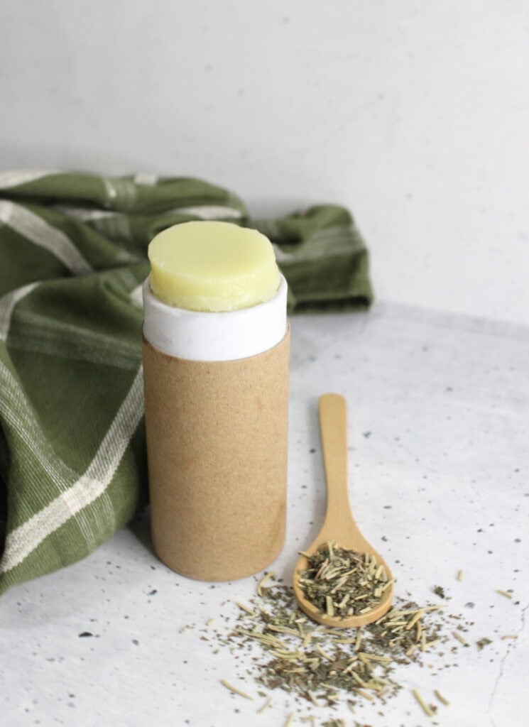 Stick of tallow-based solid lotion with herbs on table nearby.