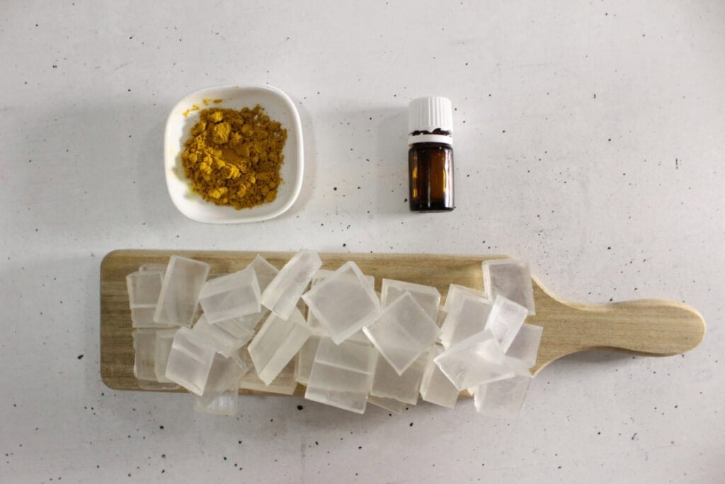 Image of tumeric soap ingredients- tumeric powder, essential oils and cubed glycerin soap base on a cutting board.