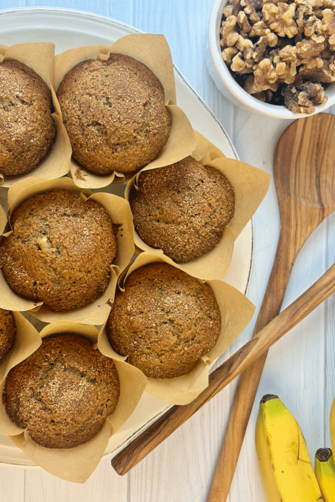 Vertical image of muffins in parchment liners on a round platter with wooden spoons and bananas nearby.