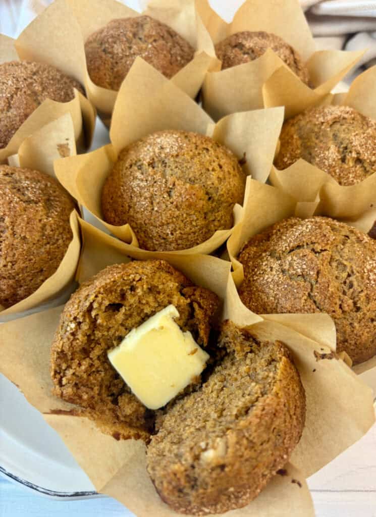Cinnamon and sugar topped muffins nestled together in individual parchment liners with one muffin opened with a slice of butter.