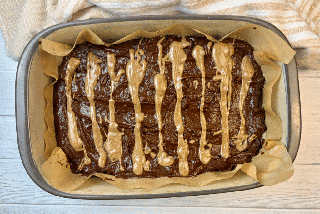 Stoneware pan with brownie batter spread in parchment paper and nut butter drizzled across it.